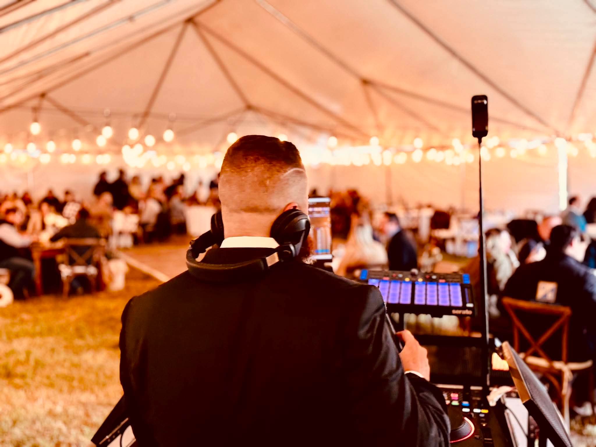 DJ Koma reading the room from behind the decks at a tented reception under warm string lights